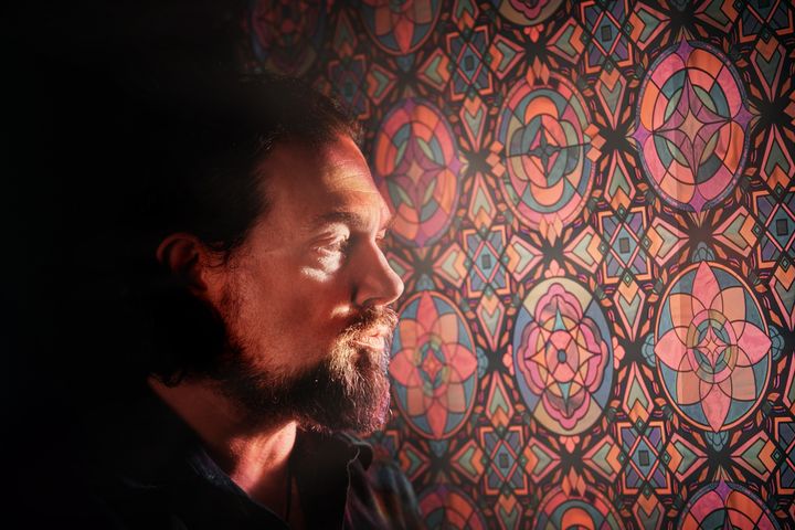 A picture of Greg Anderson looking to one side, looking at multicolored patterns on a stained glass window.