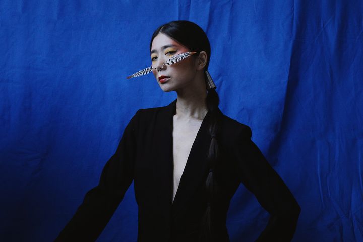 A photo of the artist Hatis Noit, dressed in black against a background of blue fabric, with feathers attached to her nostr